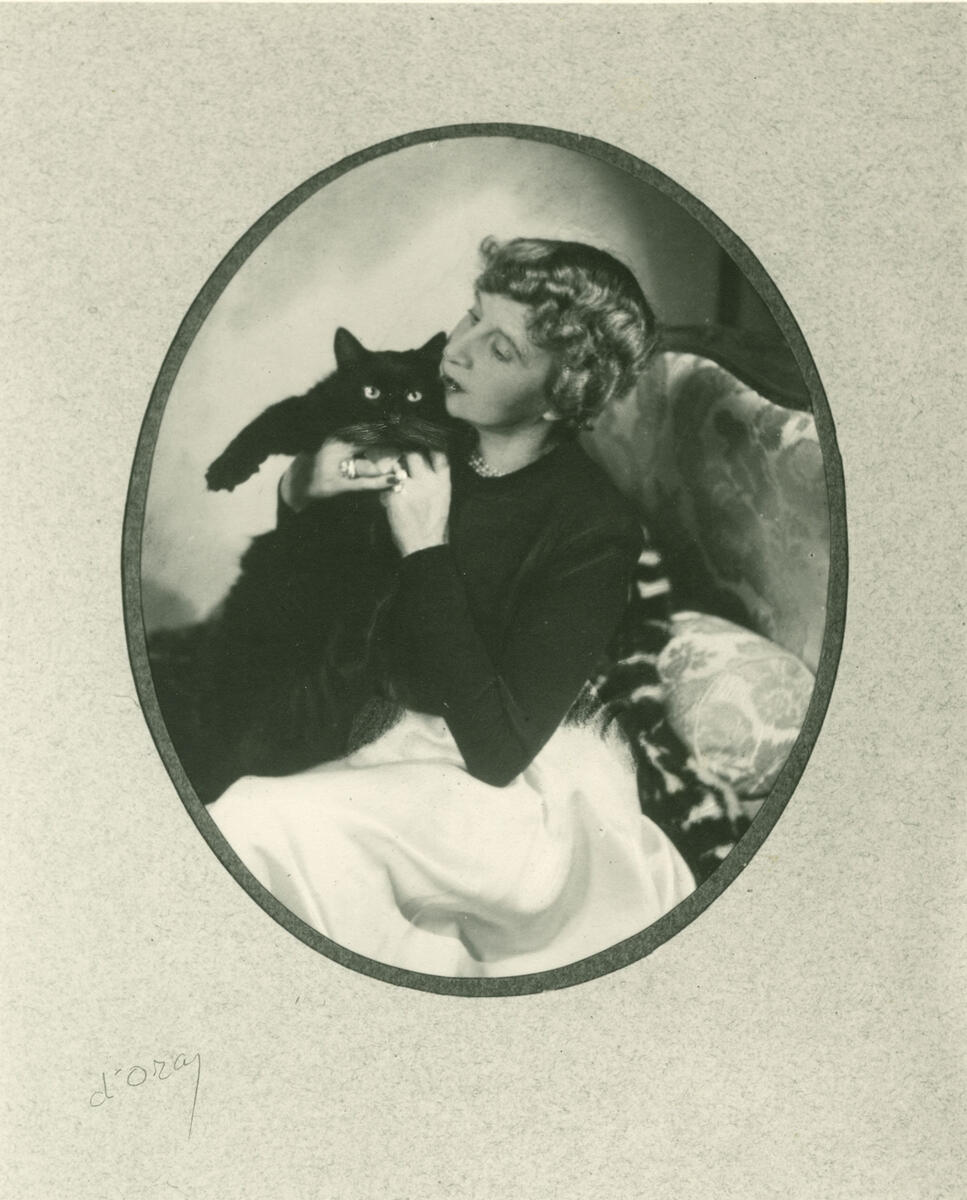 Bildet viser madamme d'Ora sittende med en sort katt i armene. Fotografiet er tatt i tisrommet 1925 til 1935 og tilhører Preus museums samling.