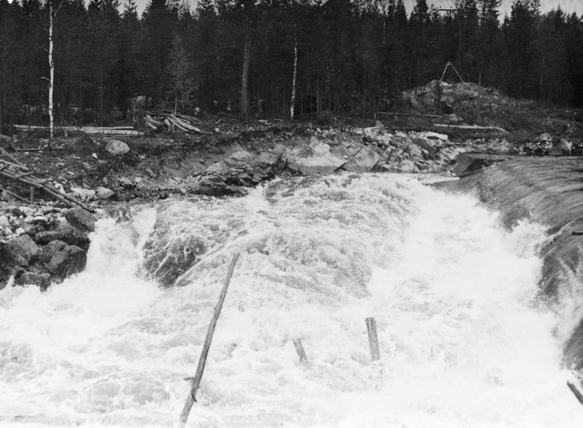 Dammen som skulle stuve opp vann til Kvernfallet kraftverk i elva Søndre Osa i Åmot kommune i Hedmark, fotografert våren 1936, like etter at anleggsarbeidet var ferdig. Her renner vannet over en avrundet terskel, utført i armert betong. Denne konstruksjonen er plassert på tvers av elveløpet, like framfor ei oppoverskrånende bergflate, som leder vannet fra nedfallssona mot den søndre elvebredden, før det føres videre vestover i det gamle elveløpet. Til venstre i bildet ser vi den nedre enden av et trestillas, et skråplan med skinnegang for traller som ble brukt under anleggsarbeidet.

Litt mer informasjon om kraftutbygginga i Søndre Osa, og om anlegget ved Kvernfallet spesielt, finnes under fanen «Opplysninger».