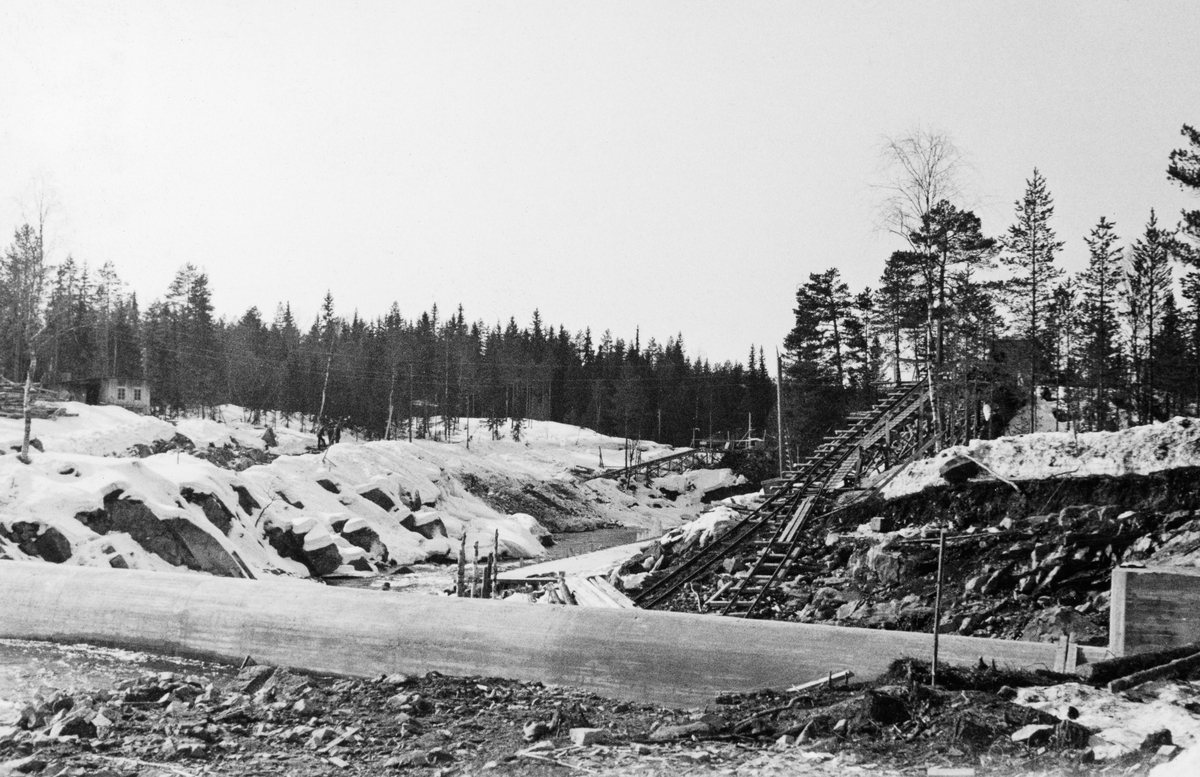 Fra bygginga av kraftversksdammen ved Kvernfallet i elva Søndre Osa i Åmot kommune i Hedmark i mars 1936, altså i en periode da vannføringa i elva var minimal. Fotografiet er tatt i medstrøms retning, med den avrundete damterskelen på tvers av elveløpet i forgrunnen. På begge sider av elveløpet ser vi også stillaser, skråplan med skinneganger for traller, som ble brukt under anleggsarbeidet,

Litt informasjon om kraftutbygginga i Søndre Osa finnes under fanen «Opplysninger».