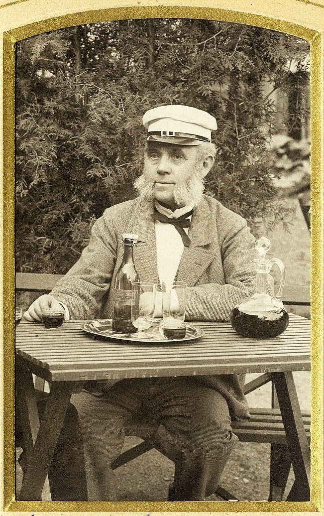 En äldre man i polisonger, klädd i ljus kostym och dito skärmmössa, sitter vid ett trädgårdsbord med en karaff och ett glas m.m. framför sig.
Knäbild, halvprofil.