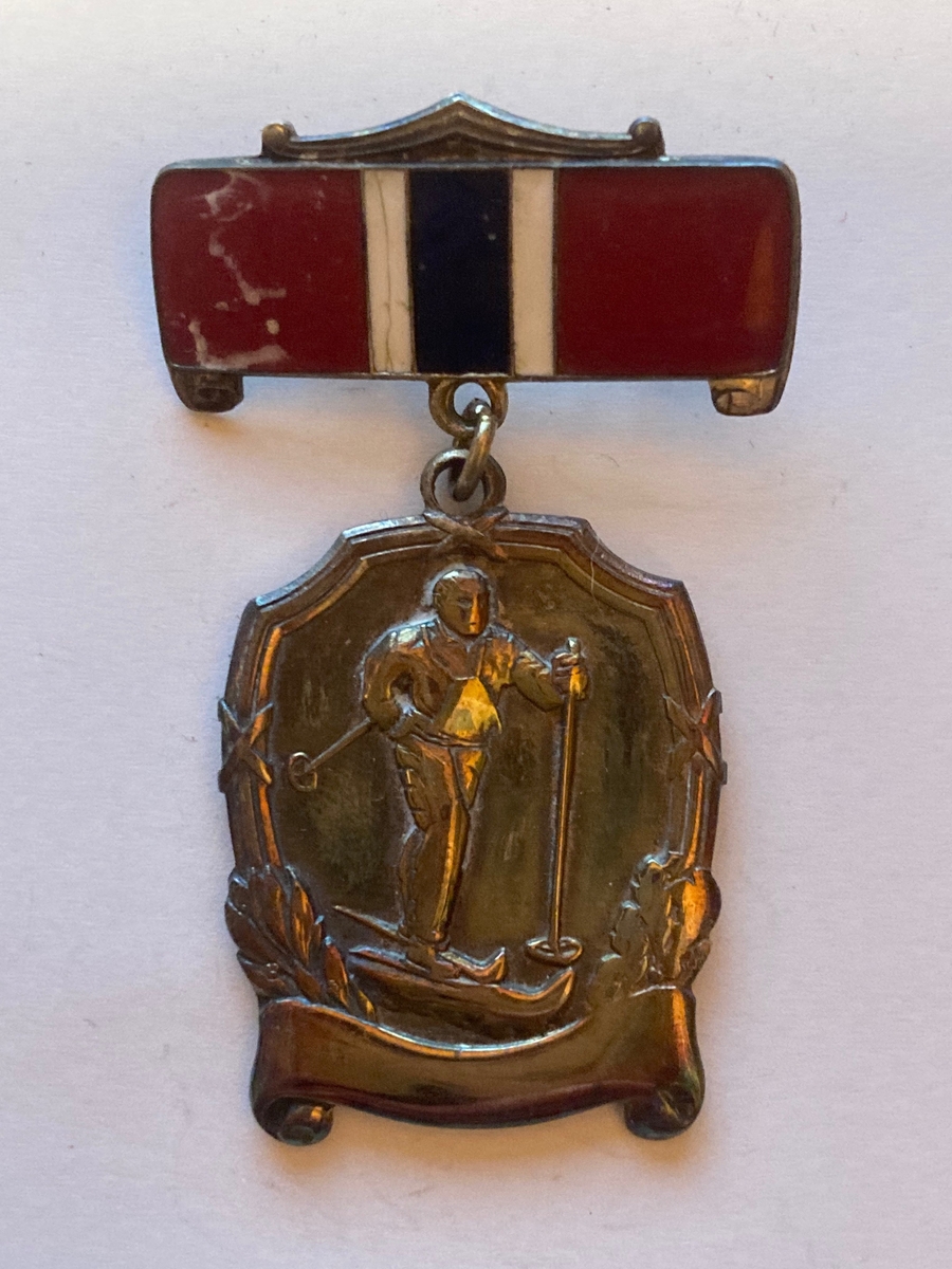 Medalje i sølv festet til en plate med nål på baksiden. Motivet på forsiden er av en skiløper.