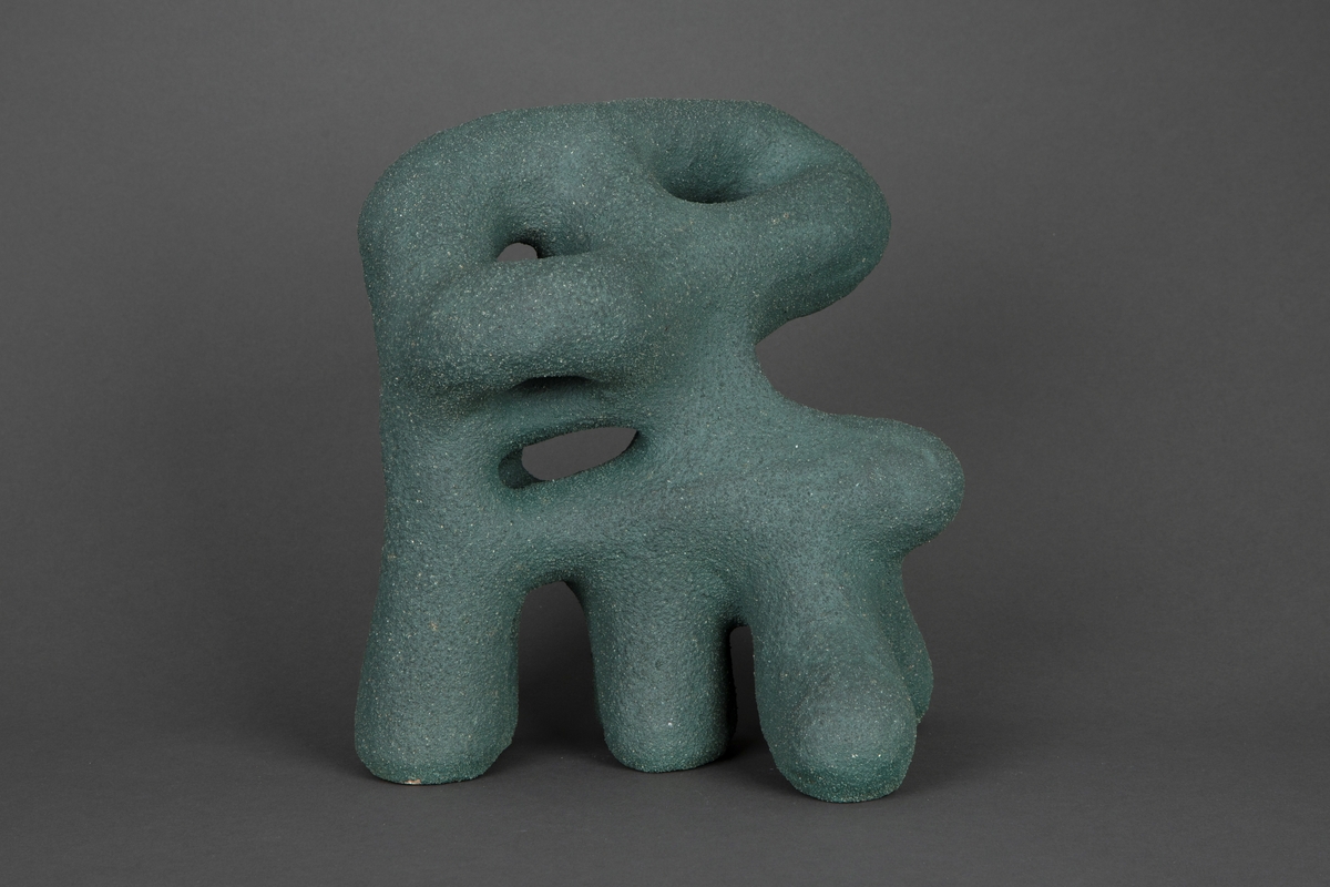 Organisk skulptur i modellert steingods. Blågrønn overflate, grovt og kornete gods. Abstrakt form, med hint til noe figurativt, som et dyr på fire bein.