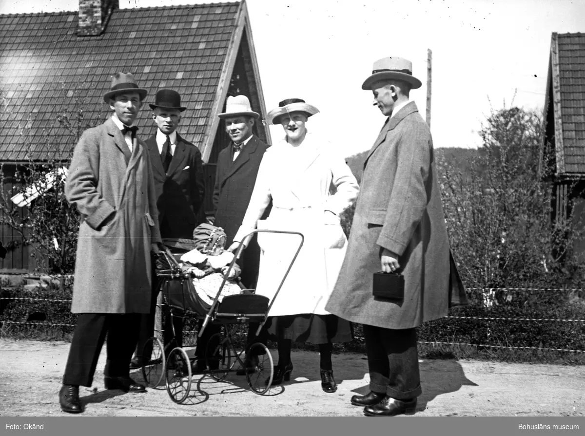 Fem vuxna och ett barn i barnvagn ståendes i vägkanten framför några hus.