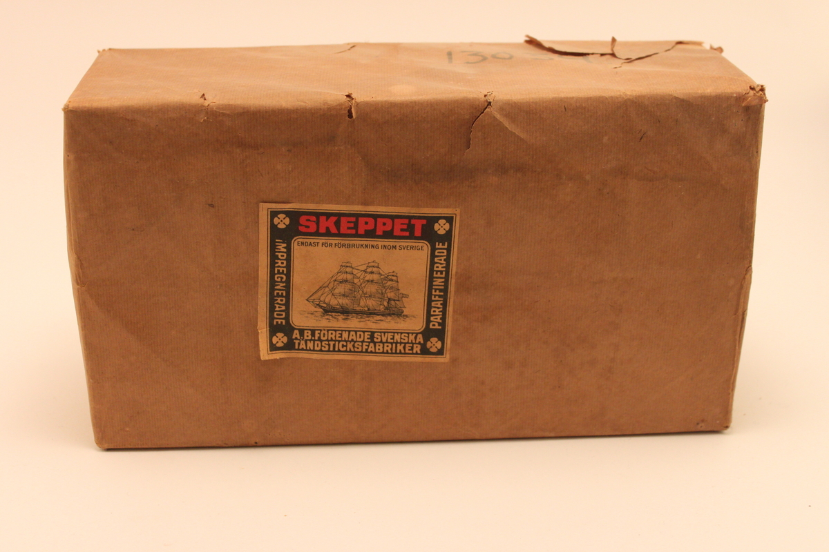 Förpackning av brunpapper med etikett. På etiketten är ett tremastat skepp avbildat. Innehåller mindre förpackningar som i sin tur innehåller tändsticksaskar av märket Skeppet. 