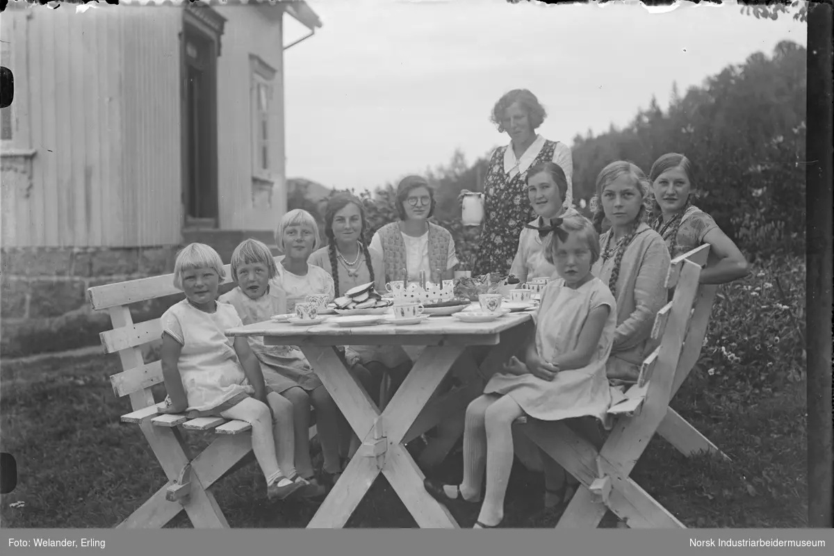 Kvinner og barn sittende rundt dekket bord. Bordet er dekket med kaffeservise og kaker. Kvinne stående ved siden av bodet med mugge i hånden.