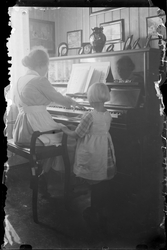 Kvinne spiller på piano i stue. Barn står ved siden av å ser