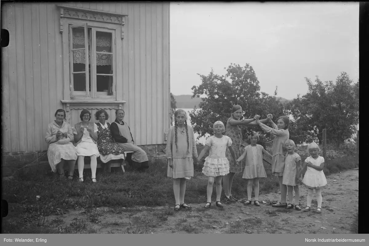 Kvinner og barn utenfor bolighus. Kvinnene sitter på benk ved husveggen, to med hekletøy i hendene. Barna leker sangleken "Bro bro brille".