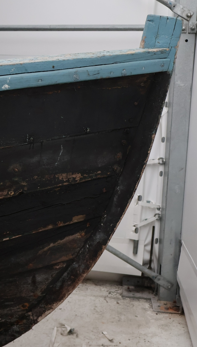 Klinkbygga storabåt med dobbel bunn, seks bord og fire keipar. Båten er svart med lyseblå ripe.