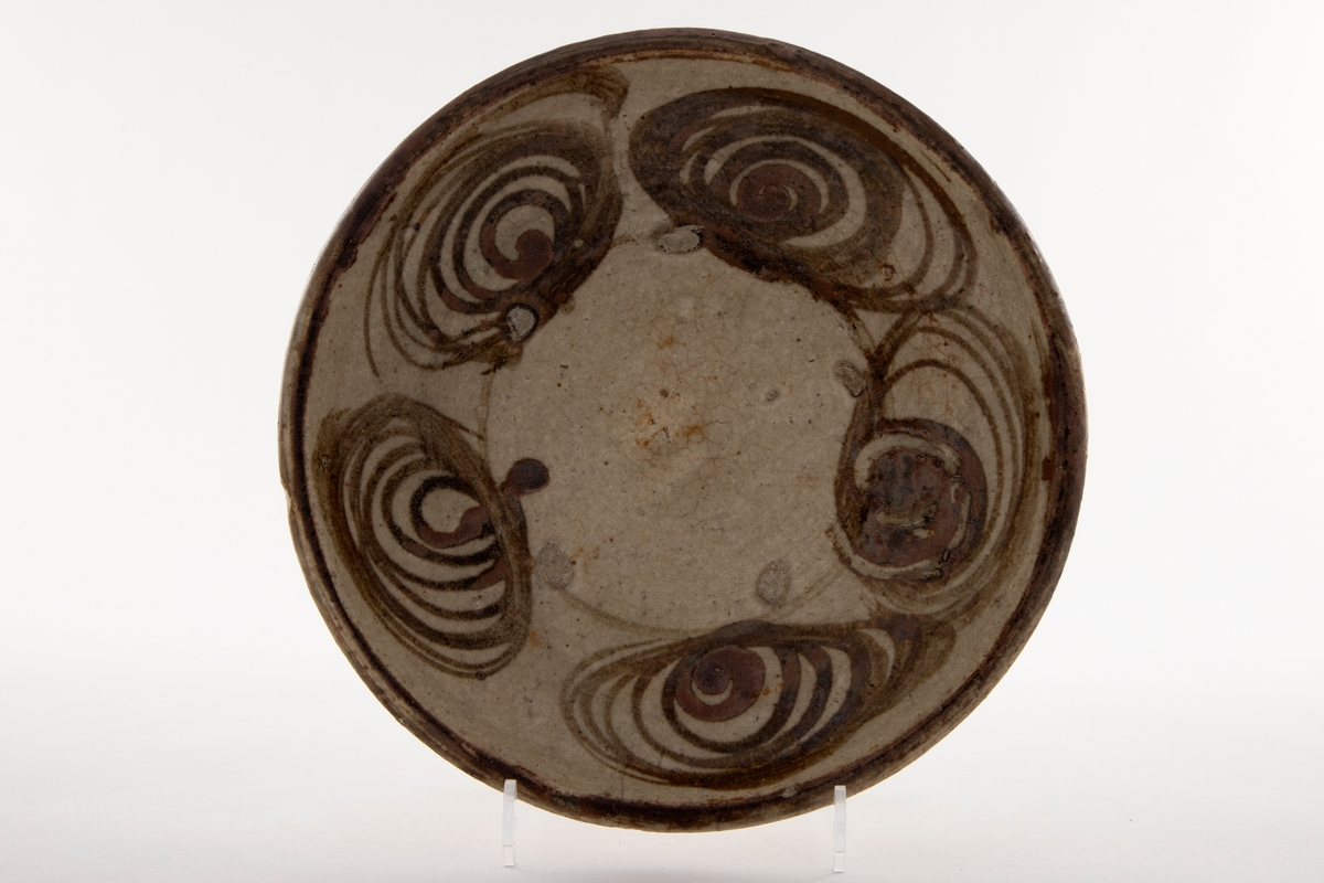 Skål eller tallerken med gulbrun glasur. Motivet viser stilliserte hesteøyne i sirkel langs kanten. Tallerkenens kant er også brun. Jevn krakelering.