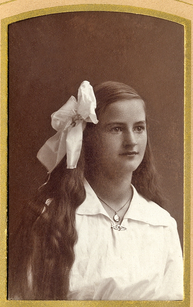 En ung okänd kvinna i vit blus och stor rosett i håret. Vid halsen syns en halskedja med medaljong.
Bröstbild, halvprofil. Ateljéfoto.