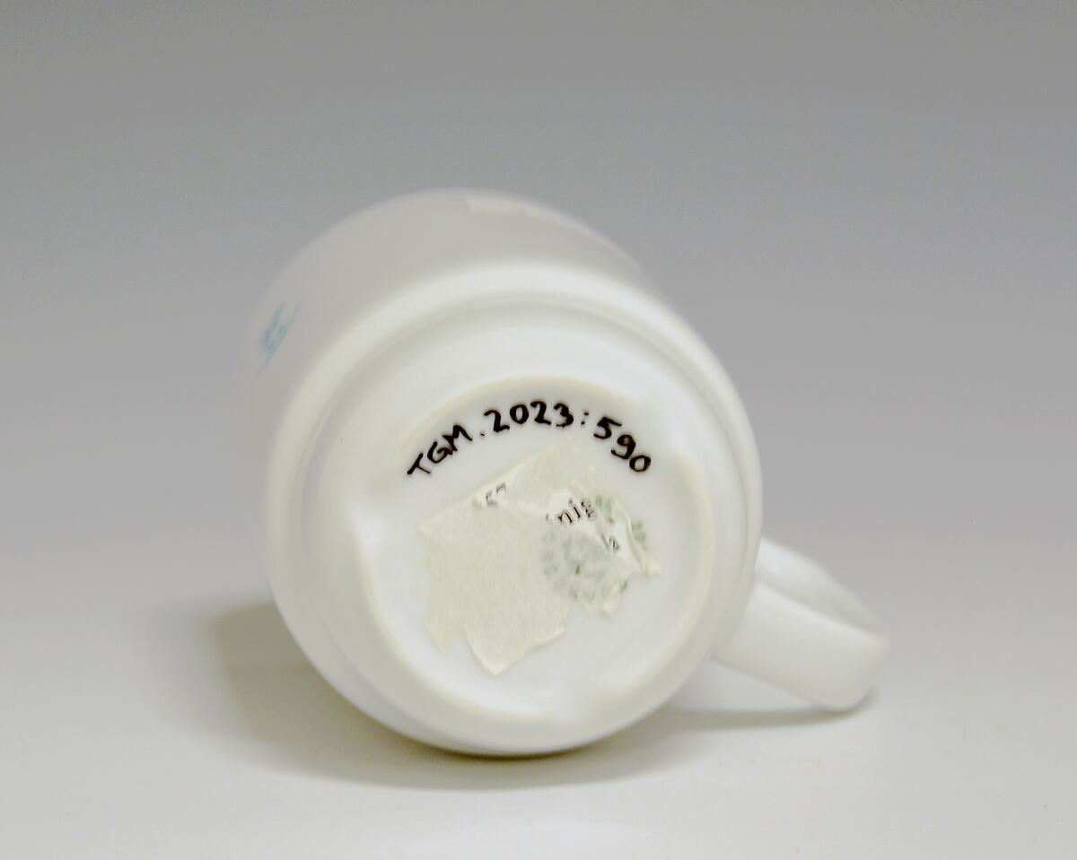 Krus av porselen med  hvit glasur. Blå logo med motiv av kokk og bokstavene "L.V.S."
Modell: Form av Tias Eckhoff