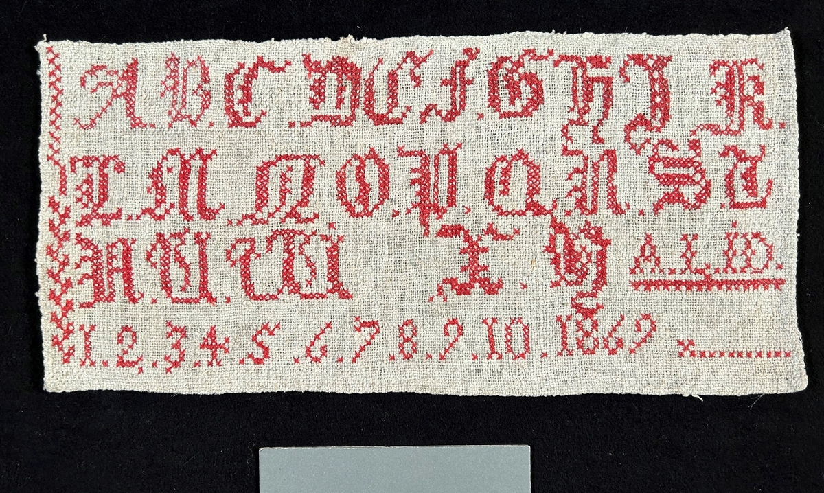 Märkduk i grått linne med korsstygnsbroderi i rött: Alfabetet A-Y, siffror 1-10 1869, samt initialerna A. L. - I. D.