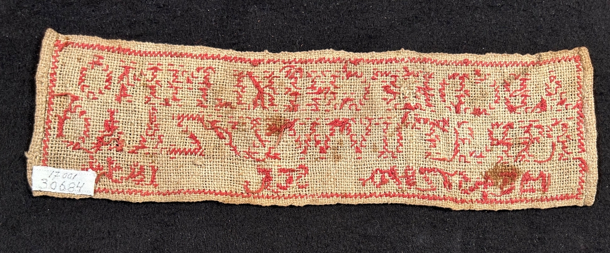 Märkduk med broderi i rött garn, alfabetet och siffror från 1-0. Tyget är av tuskaftat linne.