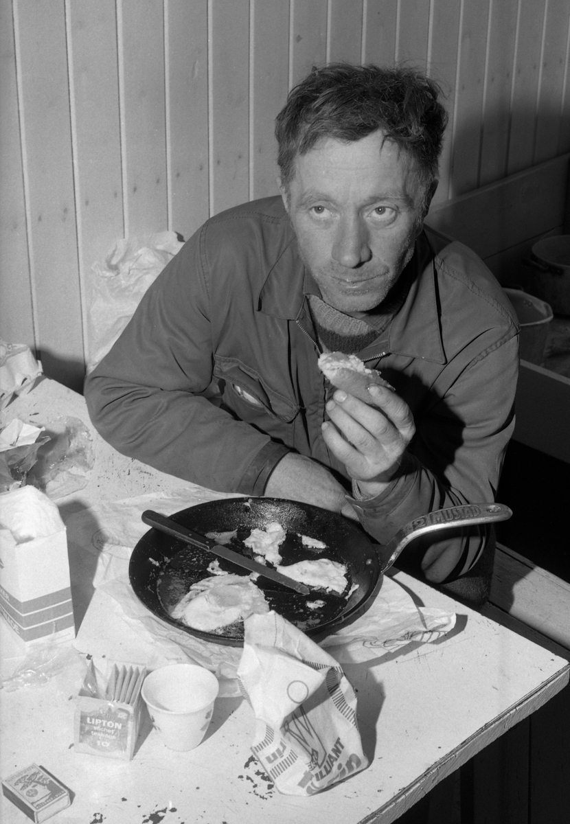 Måltid i Østlikoia i Løiten almenning (Løten allmenning) i februar 1977. Fotografiet er tatt inne i koierommet. Der satt Asmund Pedersen på en taburett under måltidet.