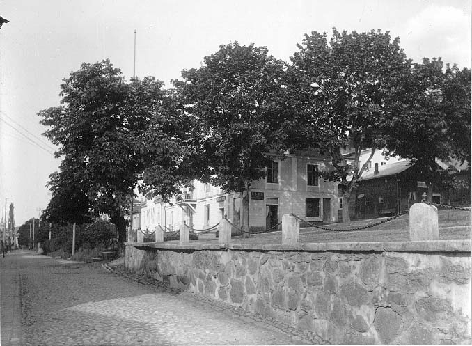 Bebyggelse vid norra änden av torget i Gränna, bakom höga träd. Torgmuren mot Brahegatan i förgrunden. Skyltar för "Hvetlanda Boden" och en garn-affär.