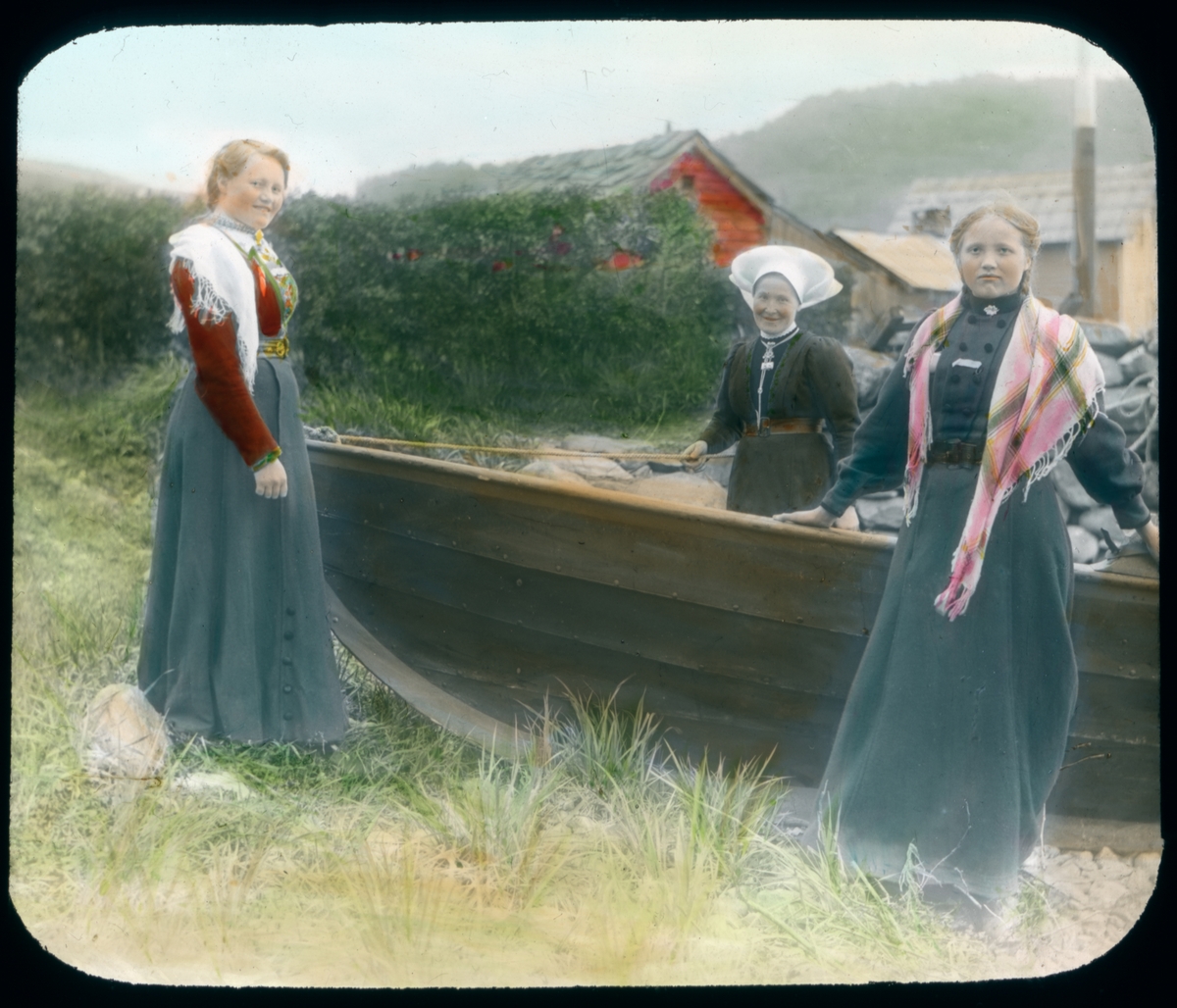 Håndkolorert dias. Tre kvinner i folkedrakter fra Hardanger står rundt en trebåt dratt opp en strand.