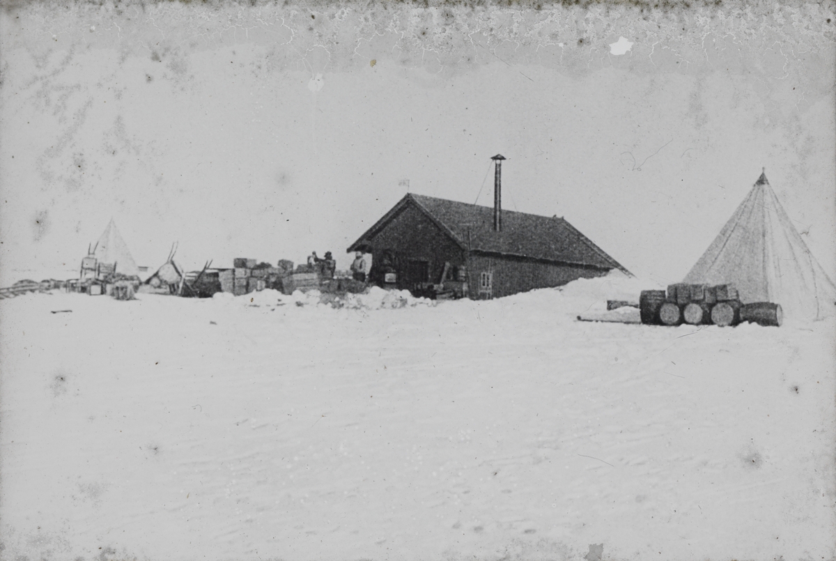 Dias. En leirplass i et snødekket landskap. Utstyr står stablet opp rundt en enkel trehytte og et telt.