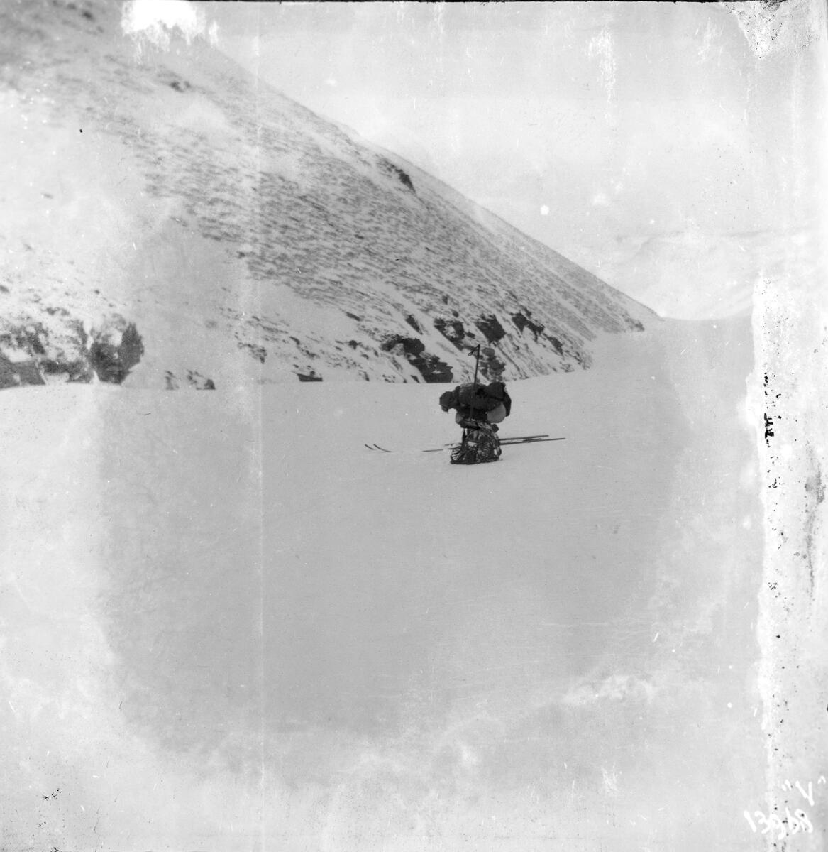 En mann sitter på huk i et snødekket landskap. Han har en sekk ved siden av skiene.