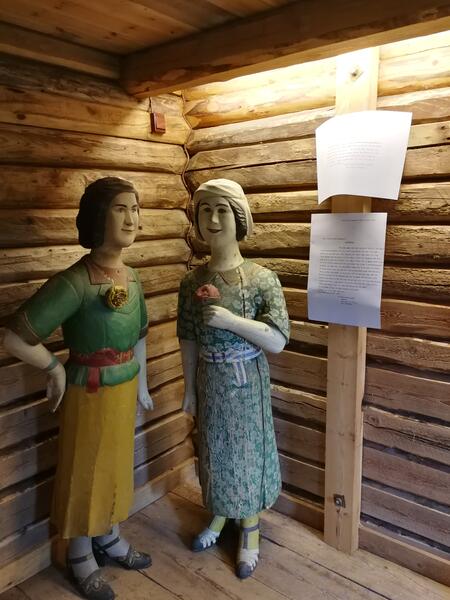 To Langsrud-figurer i utstillingen "Langsrud-kæra". Figurene viser to damer i fint tøy. De står i et rom med tømmervegger.