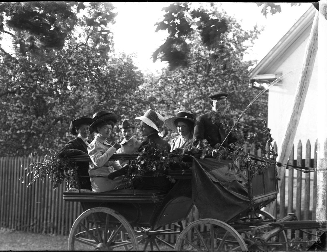 Fyra kvinnor och en man i åkvagn. En kusk på kuskbocken med en piska i handen. Vagnen står vid ett staket, på andra sidan en putsad husgavel.