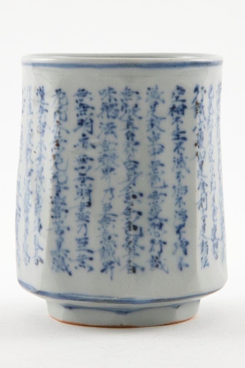 Tekopp i keramikk. Sylindrisk form med liten fot. Glasert med blå dekor som viser skrifttegn samt en blå sirkel rundt koppen ved øvre kant, nedre kant og rundt foten.