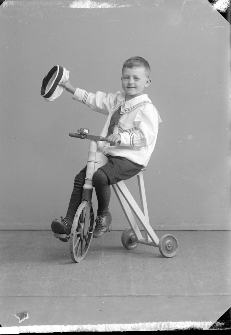 En liten pojke i sjömanskostym sitter på en trehjulig cykel och vinkar med sin keps.
