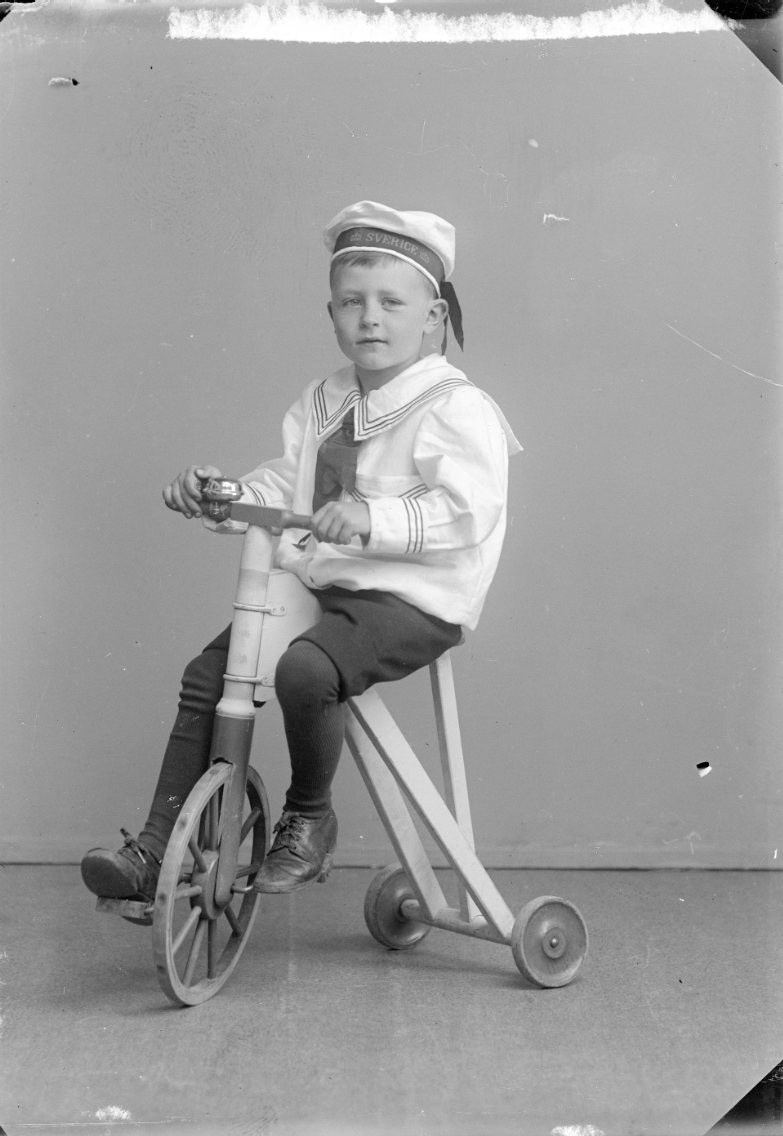 En liten pojke i sjömanskostym och keps märkt "Sverige" sitter på en trehjulig cykel.