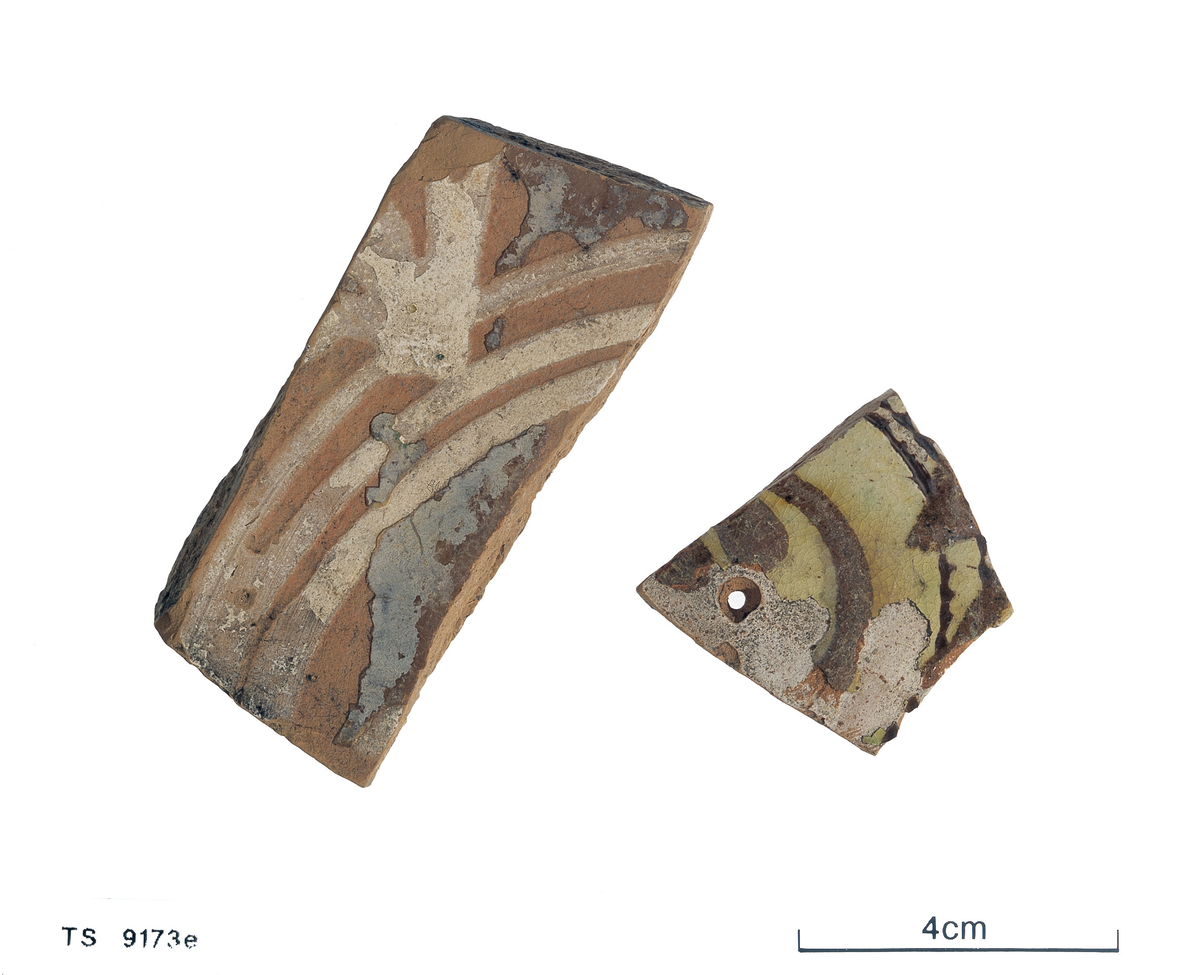 Keramikkskår m/rester av brun glasur på begge sider. Kvit begitning i stripemønster. 7,5 x 3,5 cm, 0,5 cm tykk.