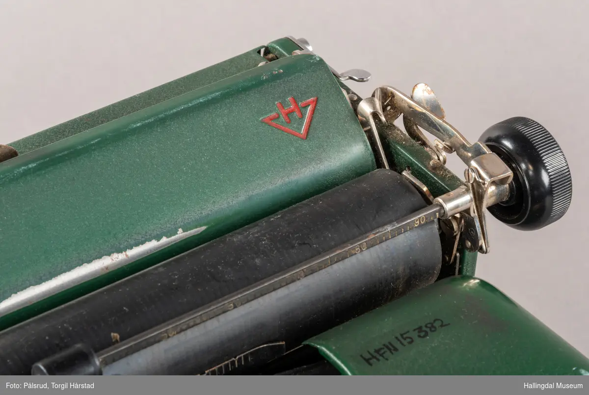 Grønn skrivemaskin av merke Halda. Den har to røde og tre gullstriper som går som en linje rundt øverste del. Noe slitasje på tastatur. 