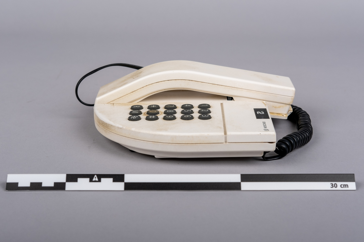 Ericsson bordtelefon.
Apparaten består av telefonrør, teelfon og spiralkabel. 
Hvit telefon med 15 svarte trykk knapper: tall (0-9), stjerne, nummertegn, funkjsonsknapper. 

Telefonen er misfarget, brune bruksmerker på telefonrør og rundt knappene, spesiell ved tallet 0.

På bunnen klistemerke med artikkel kode:
DBCN 2120700/987 R1A
ETM 9107 D. Design