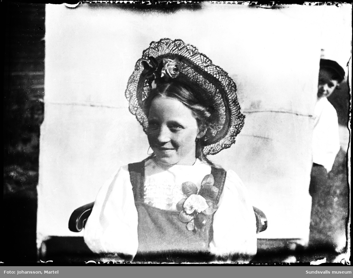 Porträttbild av en finklädd flicka med efternamnet Bergqvist. En pojke kikar fram bakom skynket.