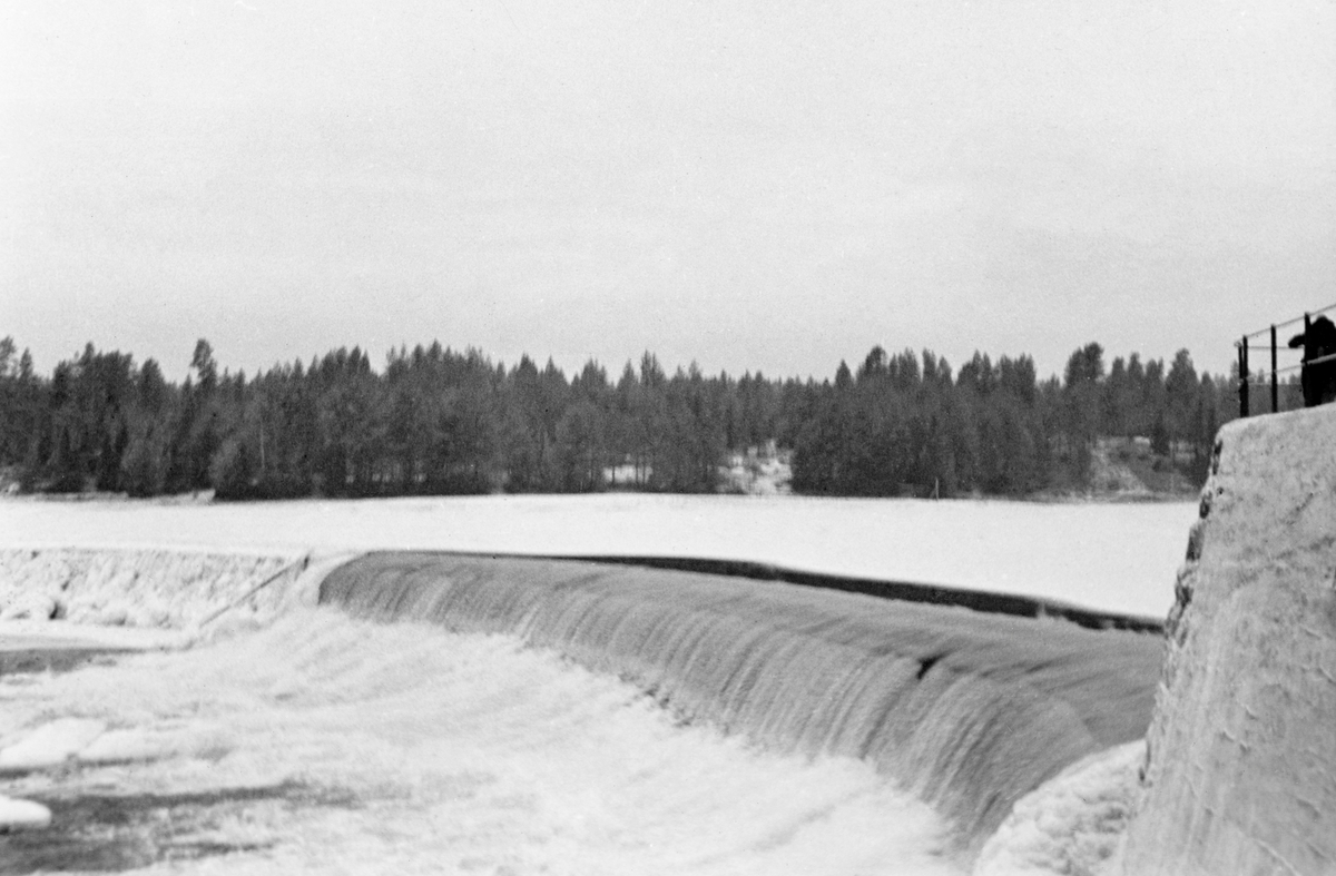 Damanlegg ved Skjefstadfossen i Glomma, Heradsbygd, Elverum, Hedmark. Fotografiet er tatt seinhøstes i 1933, fra den østre elvebredden langs den buete, 312 meter lange dammen som skulle stuve opp vann til kraftverket ved den vestre elvebredden. Det lå is og snø på de stilleflytende delene av elveløpet, men akkurat ved dammen var det en åpen råk. Det var her, i den østre delen av elveløpet, tømmeret skulle passere damkrona når fløtingssesongen begynte.

Skjefstadfossen hadde opprinnelig en fallhøyde på sju meter.  Ved bygginga av kraftverksdammen som dette fotografiet er tatt fra i 1909-1910 ble vannspeilet ovenfor Skjefstadfossen noe hevet, slik at fallhøyden ble økt til 10,5 meter.  Dette anlegget var det første store kraftverket som ble reist i den øvre delen av Glomma (nord for innsjøen Øyeren).