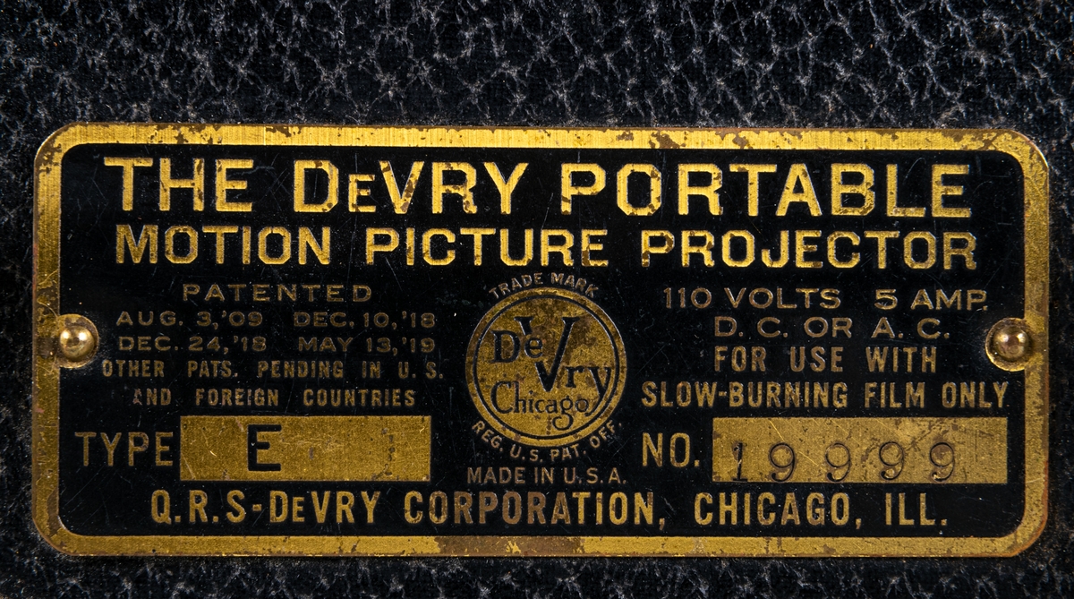 Filmprojektor i svart väska. Svart metallskylt på väskan "THE DEVRY PORTABLE MOTION PICTURE PROJECTOR". Tillverkad i Chicago, USA.
