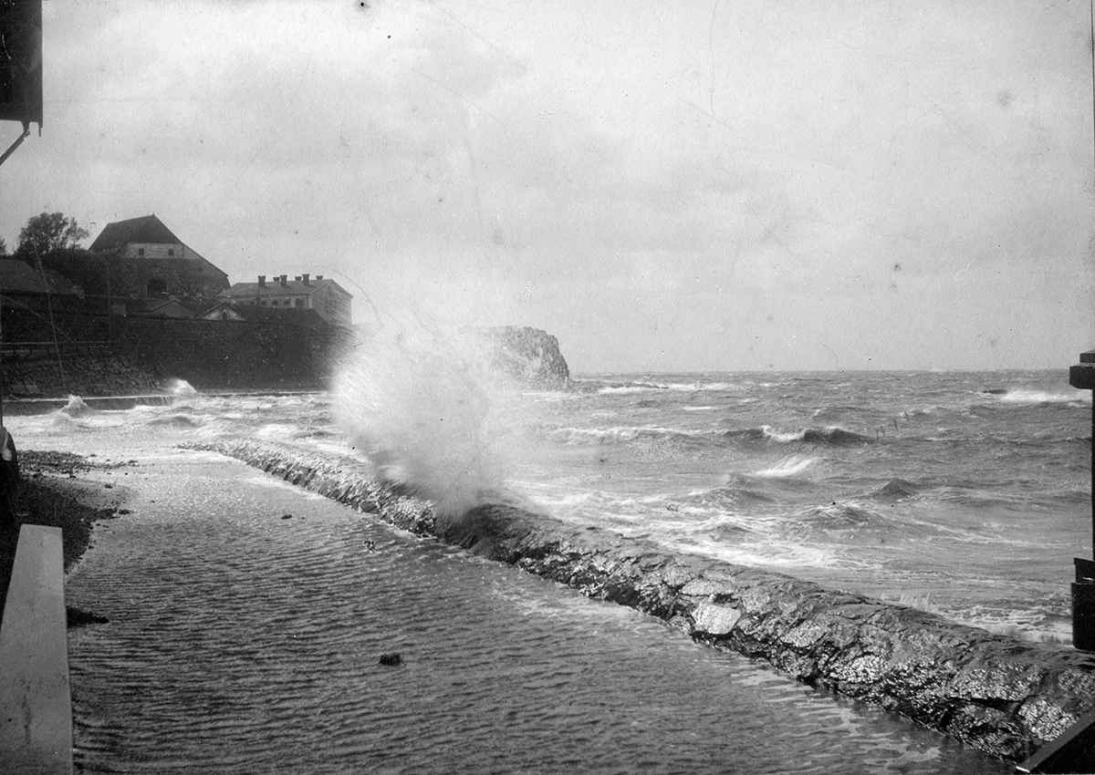 Storm, en våg slår upp över den stensatta strandkanten och vattnet går upp till varmbadhuset, som skymtar till vänster. I fonden syns fästningen.