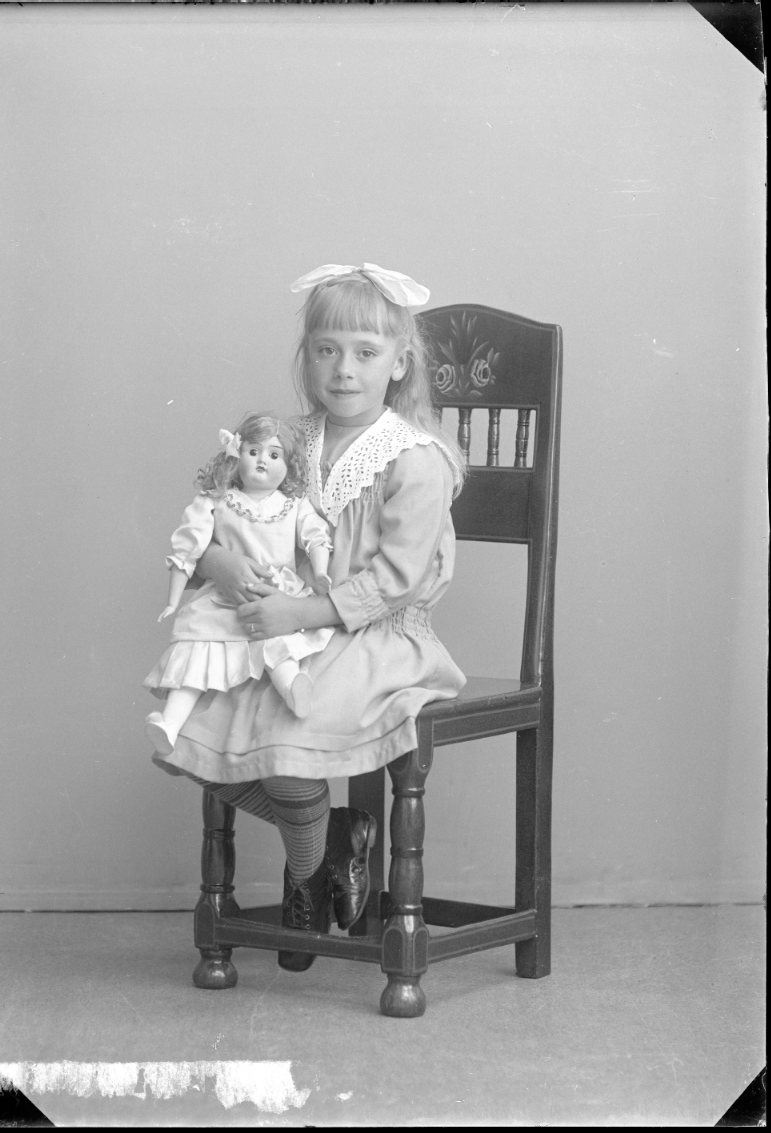 Porträtt av en flicka med rosett i håret. Hon sitter på en stol med en docka i famnen.