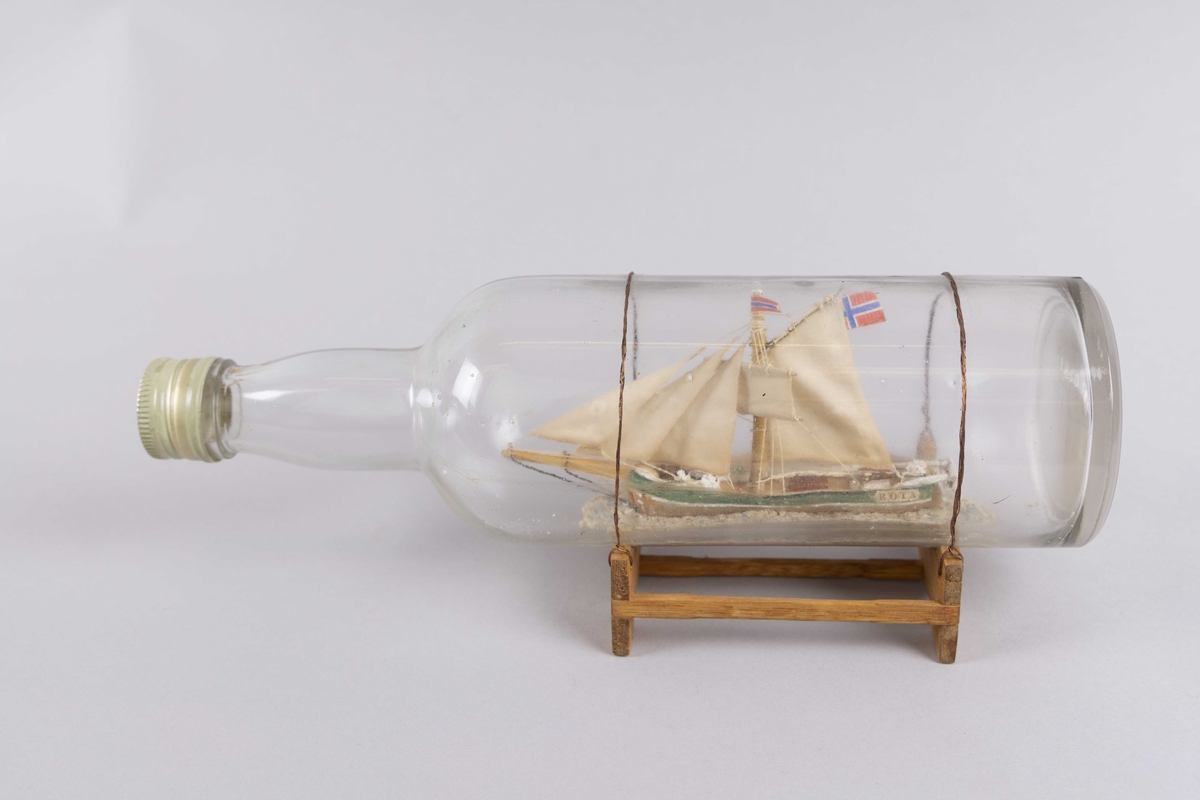 Flaskeskip av slupp ROTA med seilføring. Norsk flagg akter. Flaske montert i stativ.