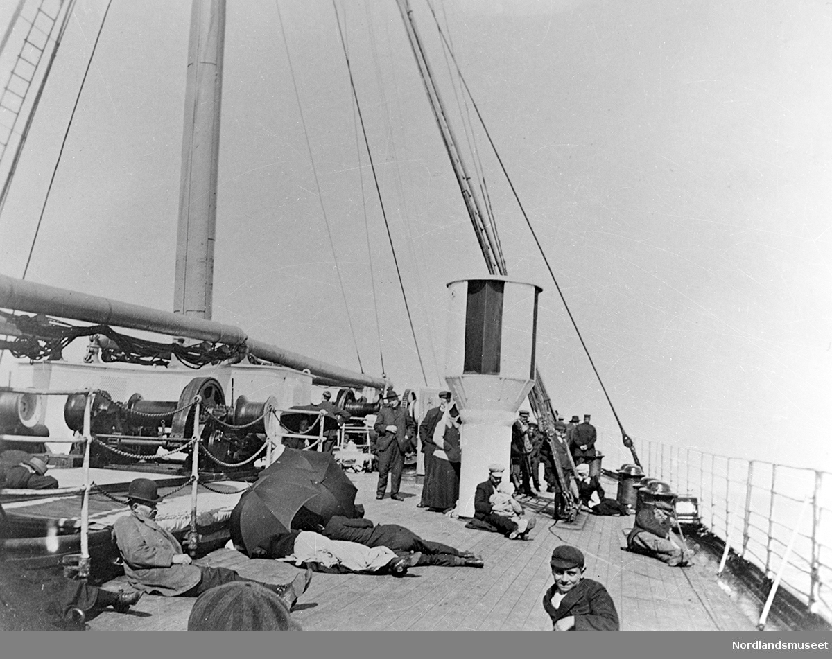 Mange mennesker som ligger og står ombord i en båt, antagelig Amerika-båten. To mennesker ligger og holder to paraplyer over seg.