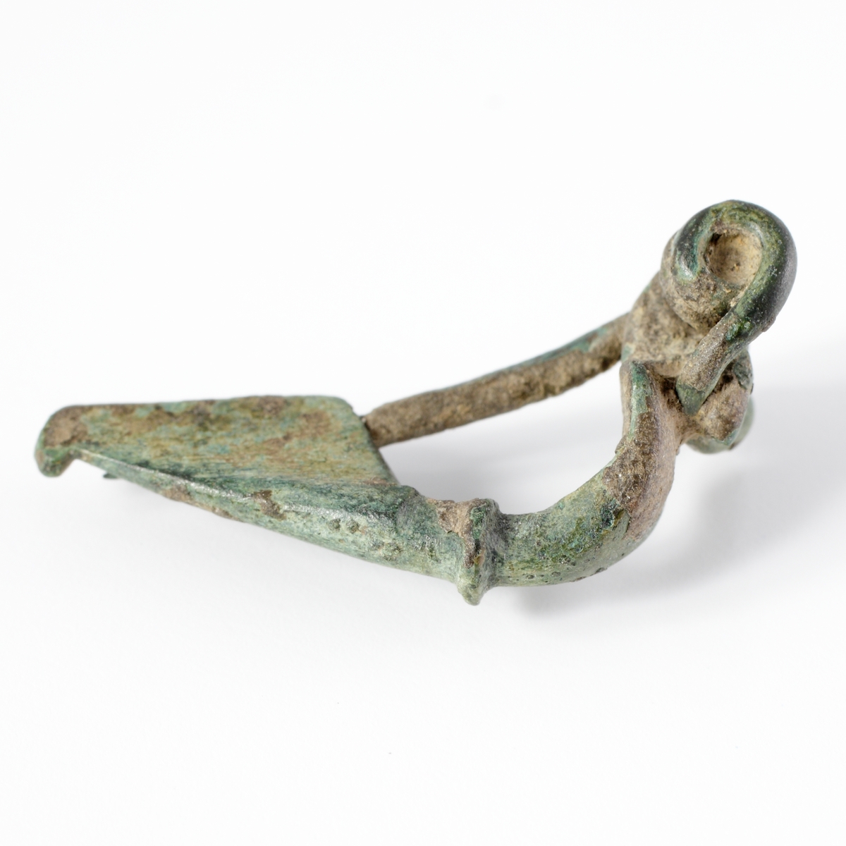 Trekantfibula av  brons med en liten vulst mellan båge och fot. Spiralfjädern fäster med en krok i änden på bågen.