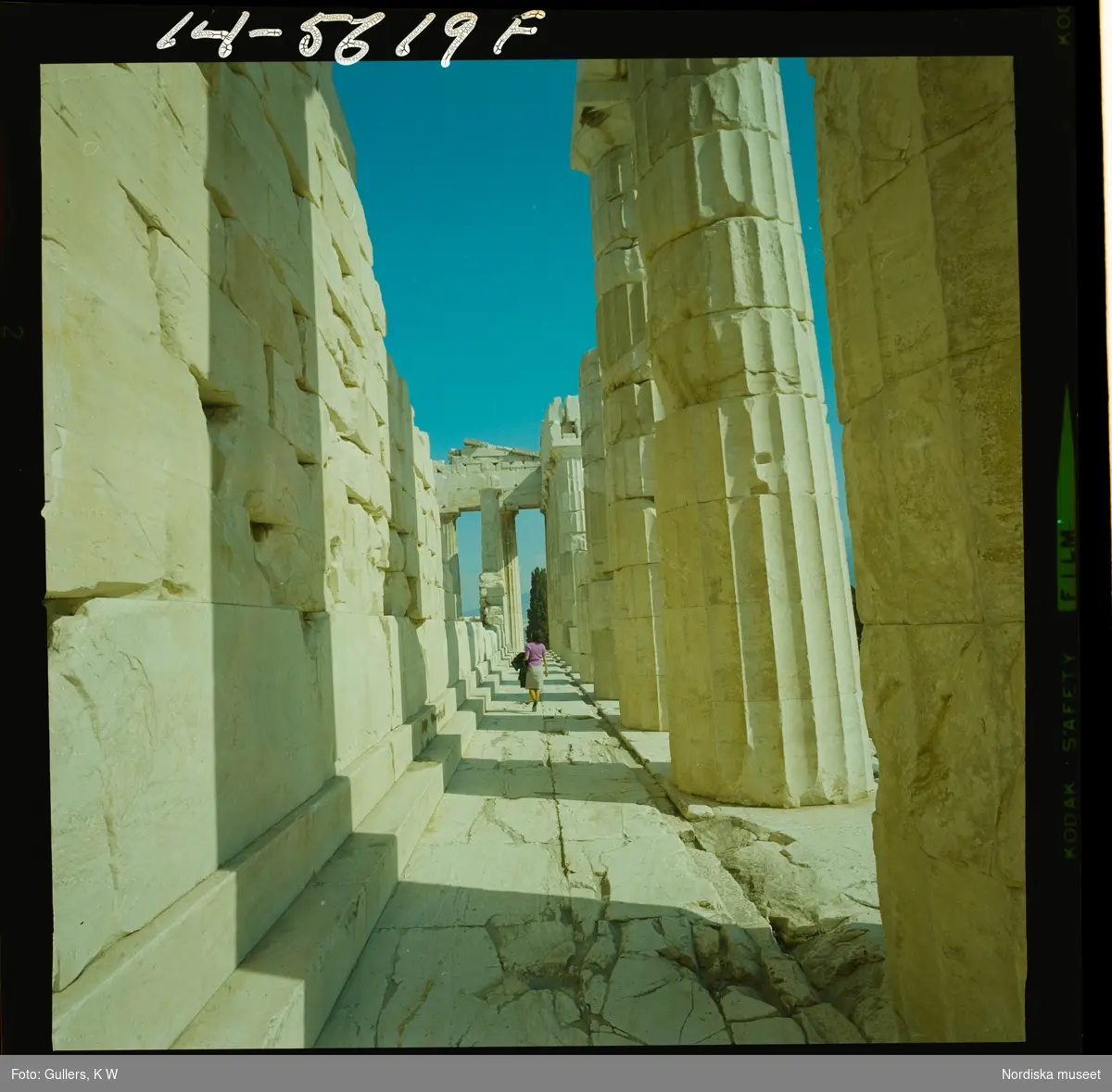 2795 Grekland Akropolis med runierna av bl.a. templen Parthenon och Erechtheion. En kvinna vandrar mellan rader av kolonner.