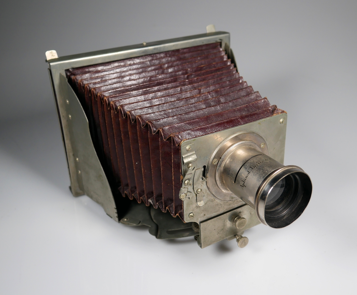 Bälgkamera med tillbehör. 
Kameran är märkt H. MADER'S Patent INVINCIBEL Model II
Modellen är den första vikbara kameramodellen och tillverkades 1888-1890. 
Väska och tillbehör är original. 

Objektivet är märkt Aplanat Invincibel. Har skåra där bländare (AM 2022-033-C) kan placeras. Bländaren har 6 valbara bländaröppningar. 

Väskan är av canvas med läderdetaljer och har ett trasigt axelband. 
AM 2022-033-A: Kamera
AM 2022-033-B: Objektiv
AM 2022-033-C: Bländare
AM 2022-033-D: Hårkors
AM 2022-033-E: Stativ
AM 2022-033-F: Väska
AM 2022-033-G:1-6: Kassetter till glasplåtar
