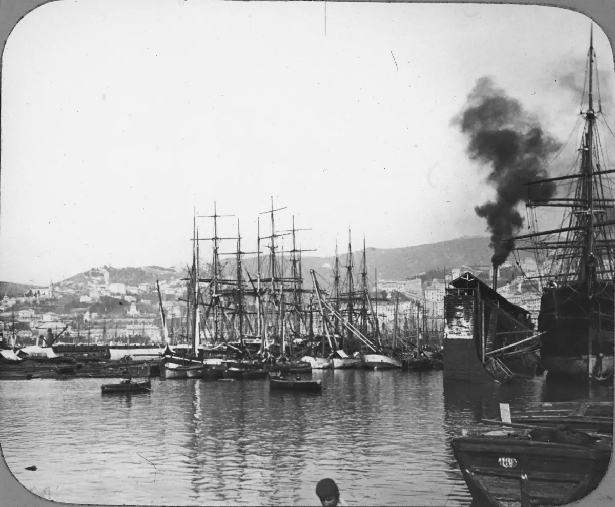 Bild från Italien föreställande hamnen Genua, "Harbour, Genoa". I hamnen finns enbart segelfartyg, men tjock svart rök, kanske från ett ångfartyg, stiger upp bakom en av de utskjutande pirerna. I bilden mitt syns två roddbåtar.
Registered G.W.W. Trade Mark.