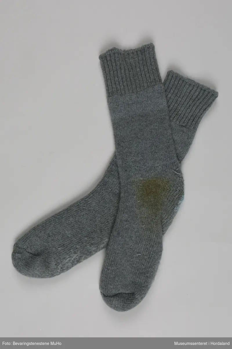 Eit par grå strikka sokkar med halvlangt brot, godt brukte. Den eine sokken er stoppa på hælen med lyseblått garn.