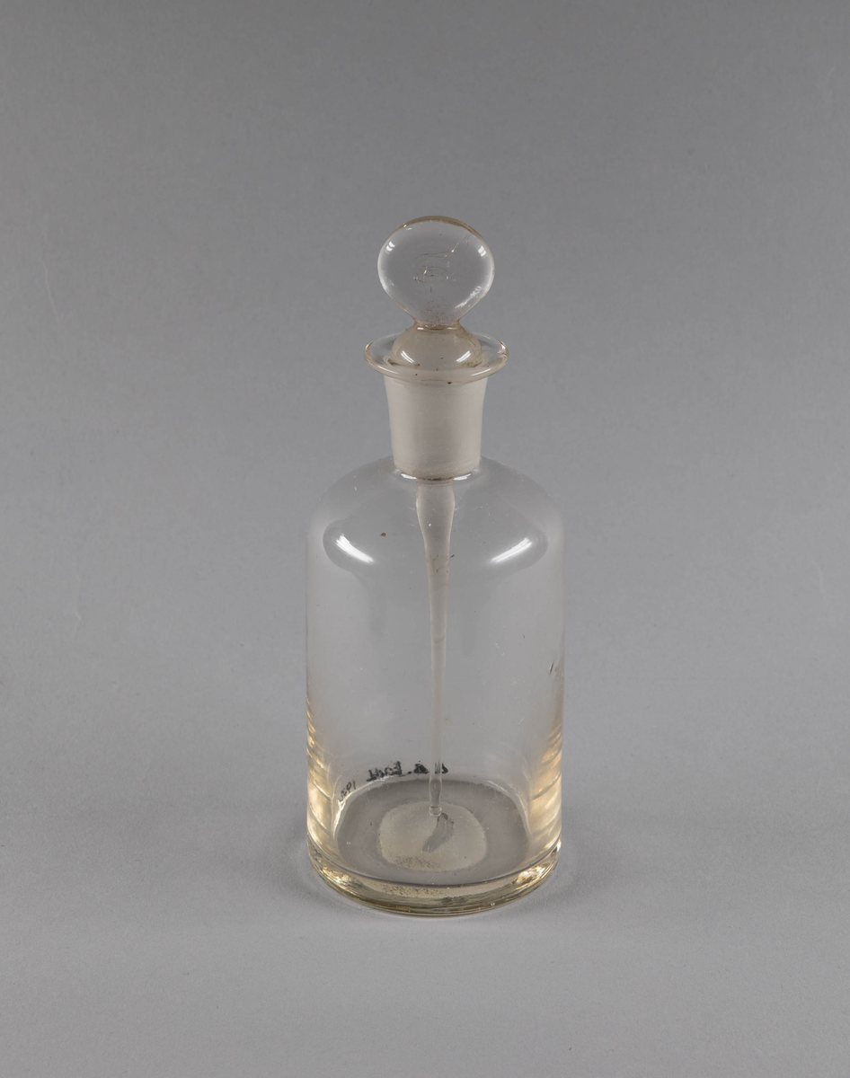 Støpt flaske og kork i glass. Flasken er sylinderformet med flat innsnevring mot rett hals. I korken er det festet et glassrør som går ned i flasken.