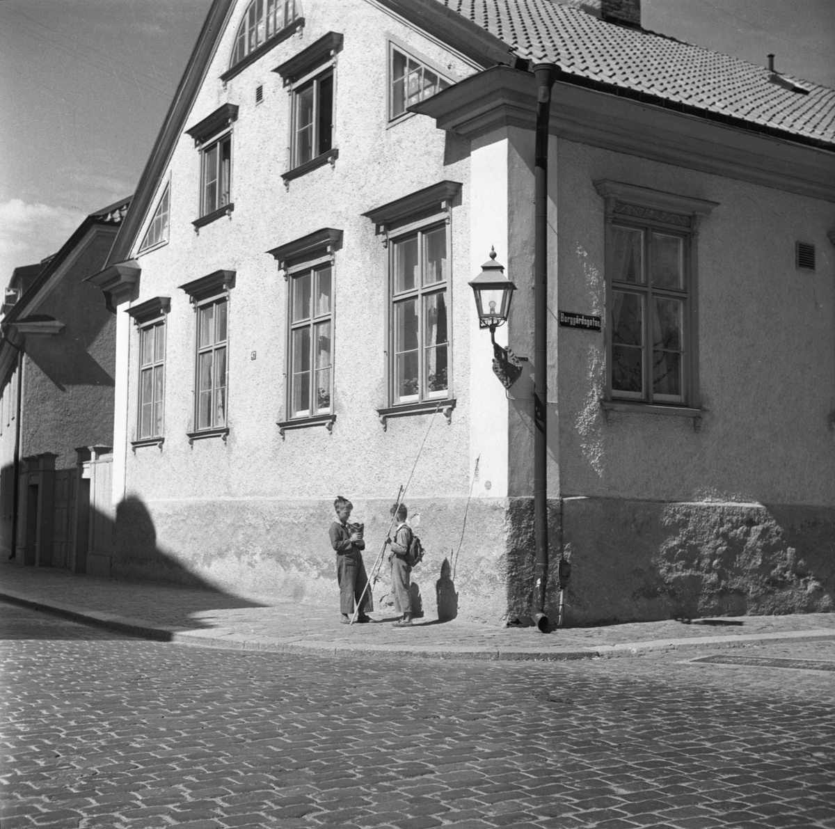 Två grabbar har stannat upp i hörnet av Storgatan-Borggårdsgatan i Linköping. Sannolikt står de i kast att promenera ned till Stångån för en stunds mete, kanske till den så kallade Mörtbryggan, ett säkert ställe för fiskelycka. Huset de står vid är stiftets domprostgård. 1950-tal.