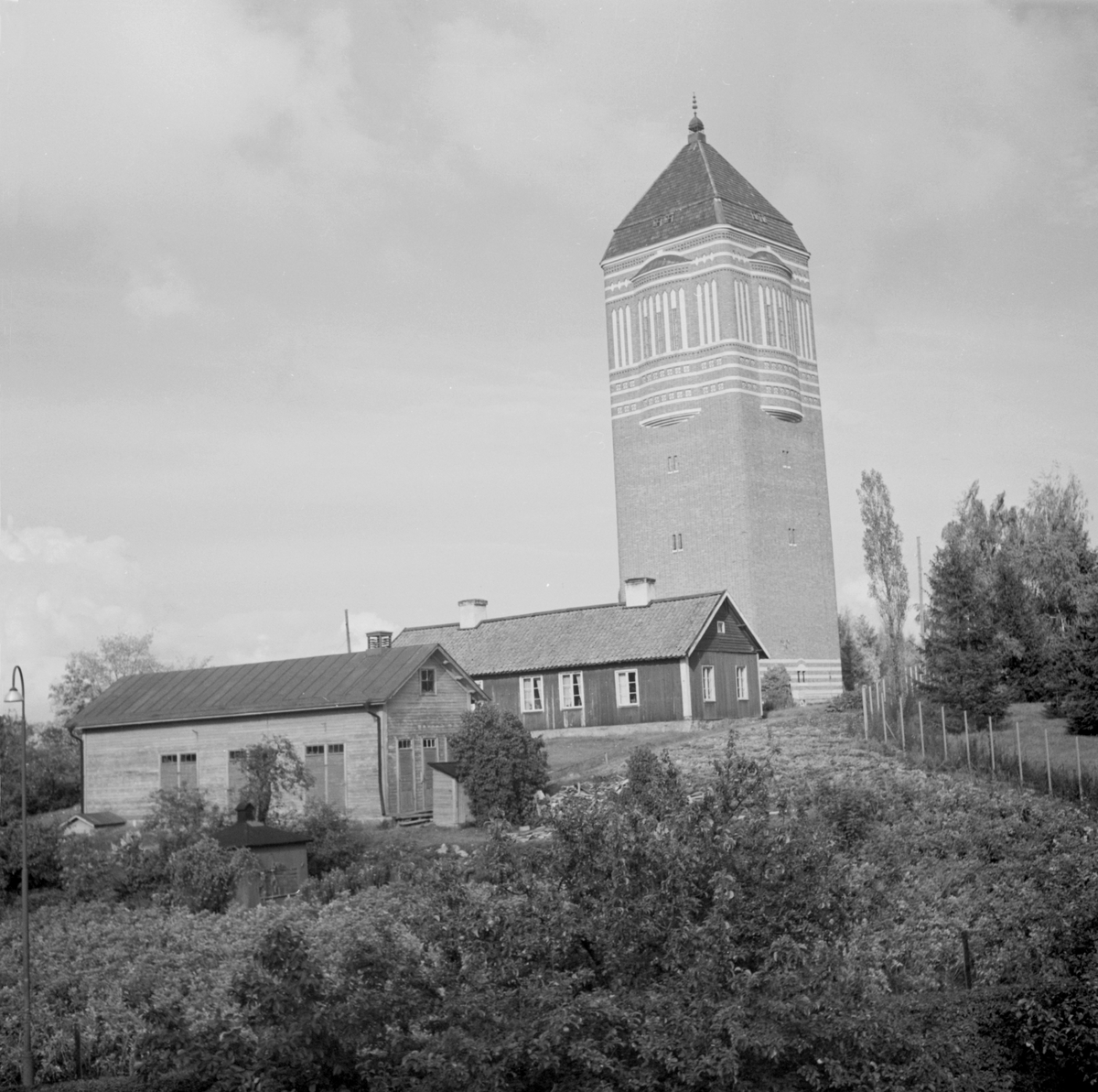 Linköpings gamla vattentorn på Kanberget. Vid fototiden omkring 1950 var anläggningen ännu i drift och hade försett stadens innevånare med rent vatten sedan 1910. För ritningarna till byggnadens monumentalt, nationalromantiska uttryck stod Axel Brunskog. I förgrunden hukar ett par enkla byggnader på ofri grund. Området hade tidigare ägts av kyrkan men den mesta av marken hade från 1910-talet styckats av till villatomter. Alldeles inpå tornet önskade dock ingen bygga, varför äldre hus stod kvar ännu in på 1950-talet.