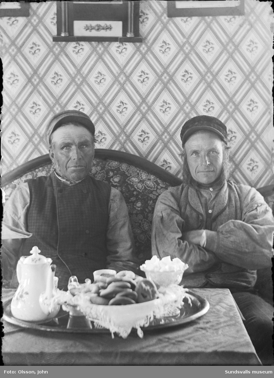 Porträttbild inomhus med fotografens bror Olof Olsson och grannen Petter, "där väst vid sjön i Ingeborgjorden" (Sunne, Värmland). På bordet framför står kaffebrickan dukad.