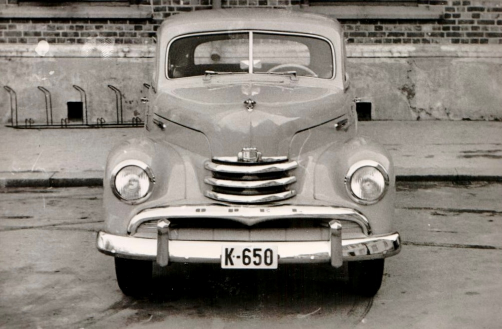 Sivil personbil av merket Opel Kapitän (årsmodell 1950-53) med registreringsnummer K-650 står parkert foran en bygning. Bilen er fotografert både forfra og fra siden.