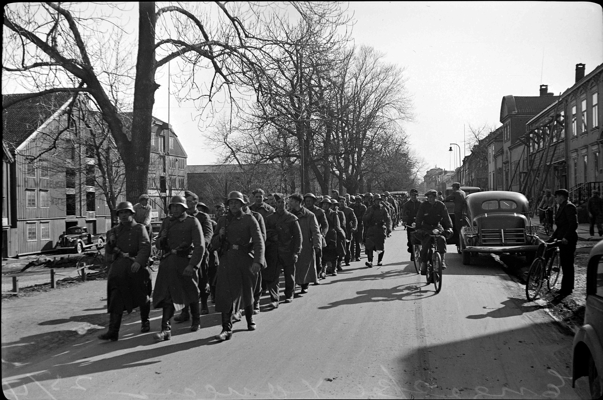 Bilde fra 2. verdenskrig. Engelske krigsfanger marsjeres ned Kjøpmannsgata. Bevæpnede tyske soldater går fremst og ved siden av rekken med engelske soldater. Til venste vises deler av bryggerekka. Til høyre er det en rekke med parkerte biler og flere tilskuere.