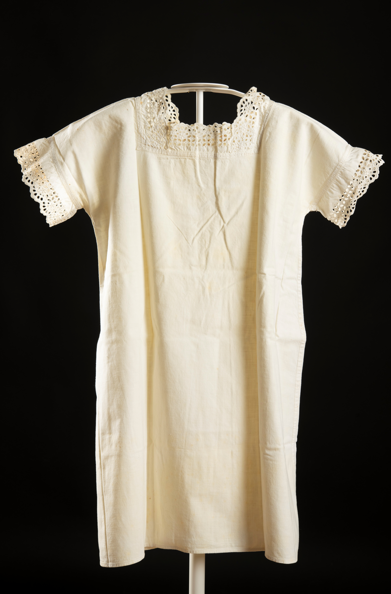 Kortärmad klänning av vitt vävt tyg. Spets runt ärm och halslinning. Bak ett 20 cm sprund med en knapp högst upp. Tyget är missfärgat på flera ställen.