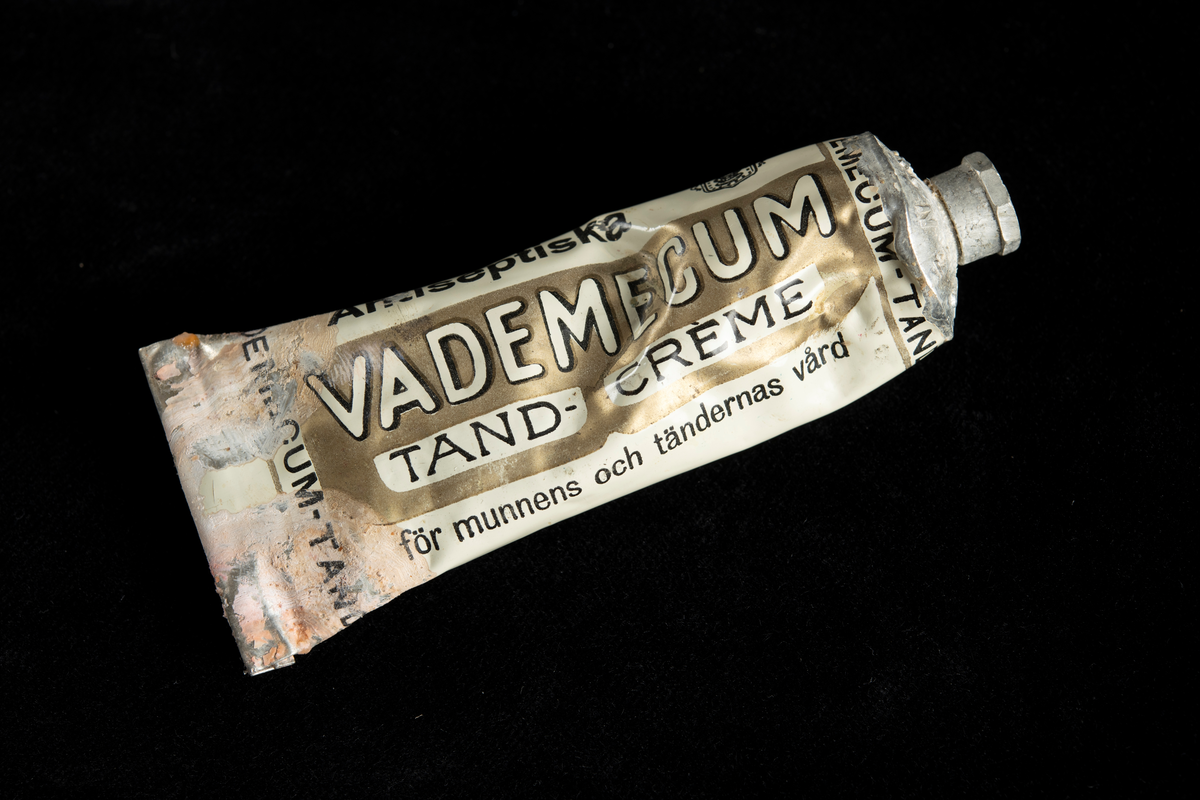 Metallförpackning innehållande tandkräm av märket Vademecum. Text i vitt och svart mot vit eller guldfärgad botten.